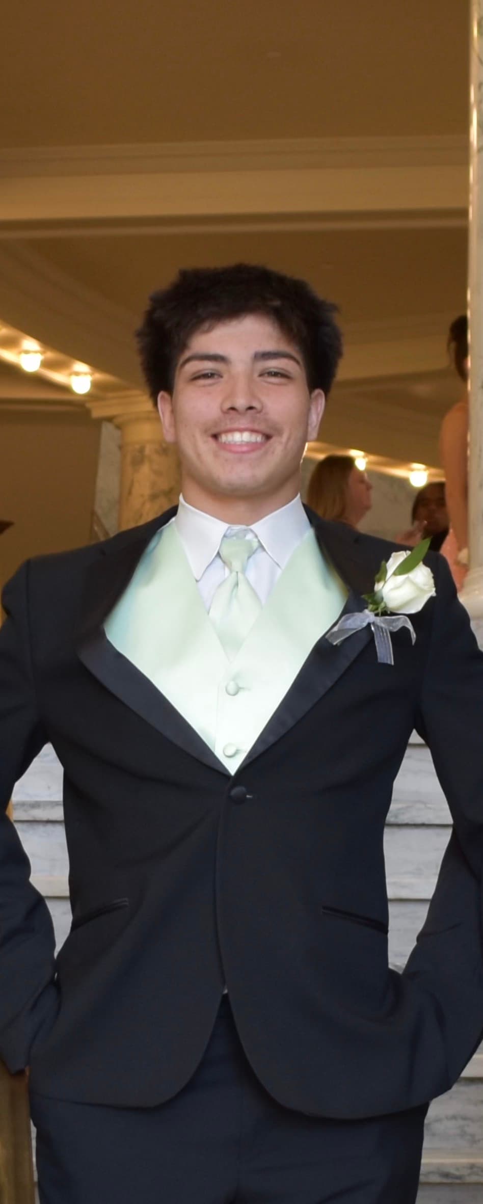 Smiling young man wearing a black tuxedo, light green vest, and a white rose boutonniere.